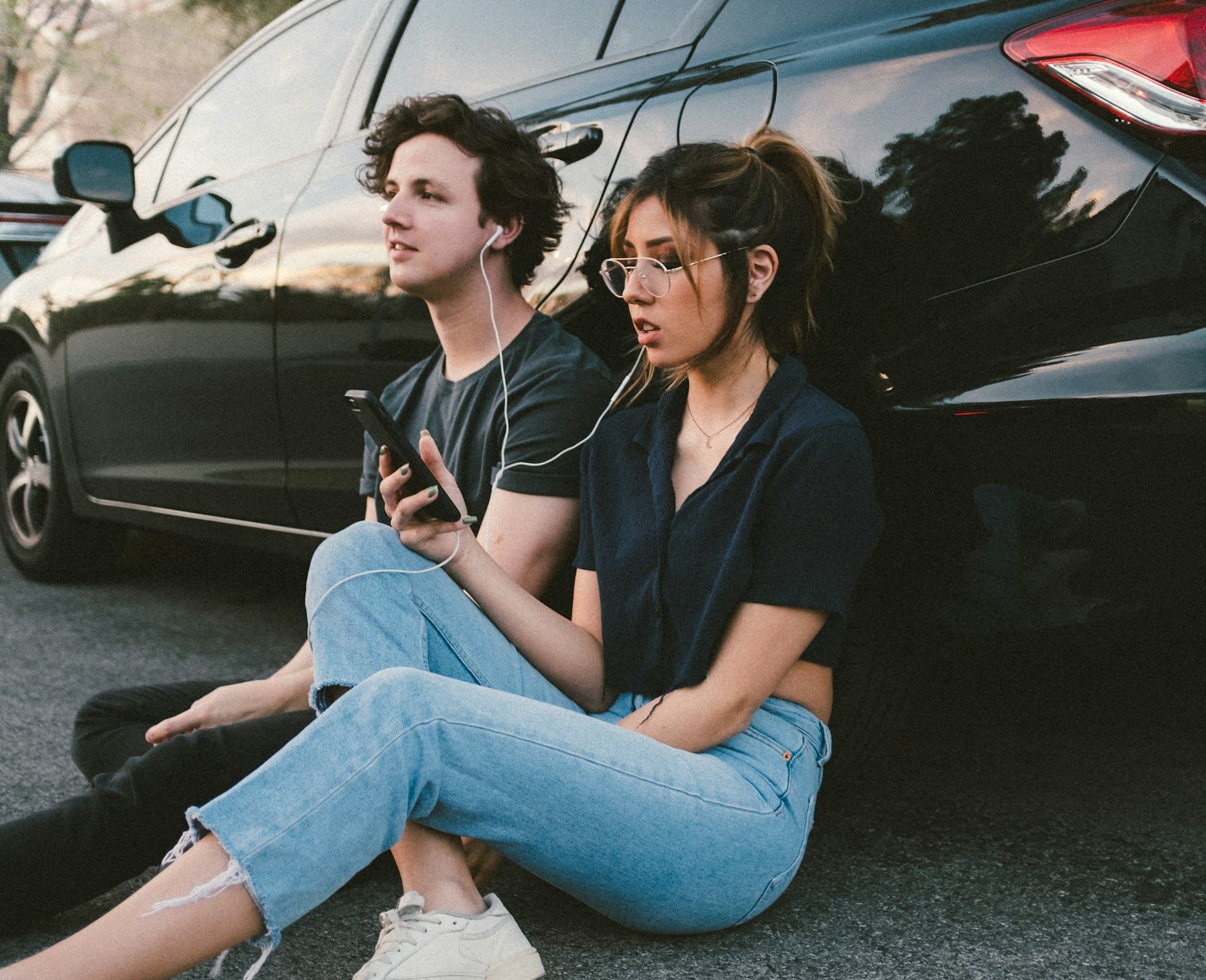 Two people sitting on the ground beside a car, sharing a pair of wired earphones from one phone